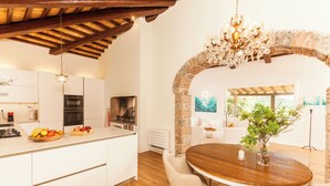 Una cucina moderna con mobili bianchi, un'isola centrale con frutta, una zona pranzo con tavolo e sedie in legno e un arco in mattoni.