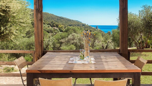 Un tavolo di legno con sedie, un vaso con fiori e vista su un paesaggio costiero.
