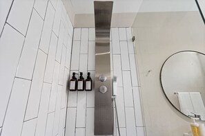 Une salle de bain moderne avec des murs recouverts de carreaux blancs, une douche dotée d’un panneau de commande et un miroir rond.