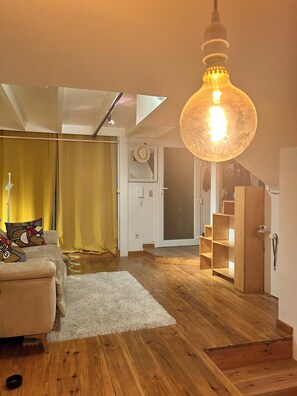 A modern living room with a sofa, a large hanging light fixture, wooden flooring, and a staircase leading to an upper level. - AT THE FOOT OF THE ARENES: AN ARTIST'S APARTMENT (Arles)