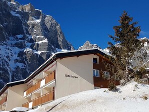 Ein Gebäude mit der Aufschrift 'Gitzifurka' an der Fassade, umgeben von schneebedeckten Bergen und einem klaren, blauen Himmel.