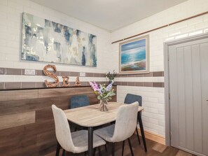 A dining area with a wooden table, two chairs, a vase with flowers, and a wall with a 'SEA' sign and abstract art. - Shoreline Apartment No.3 (Newquay)