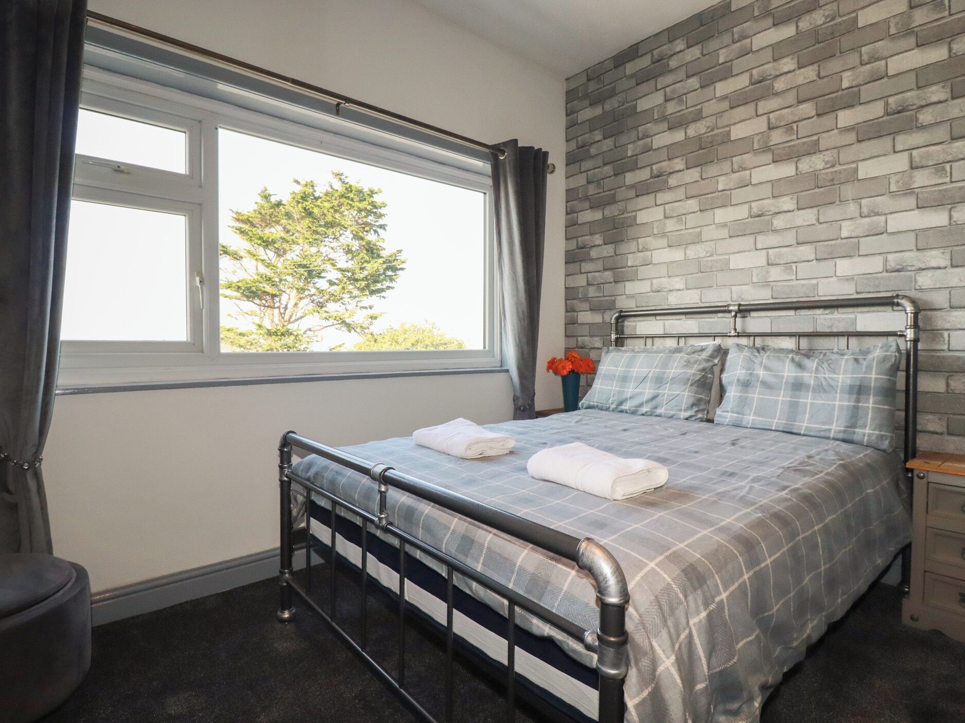 A bedroom with a metal bed frame, a window with curtains, a brick wall, and a view of trees outside.