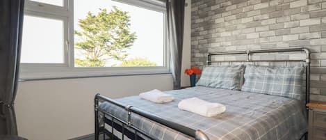 Une chambre avec un lit en métal, une fenêtre avec des rideaux, un mur en briques et une vue sur les arbres à l’extérieur.