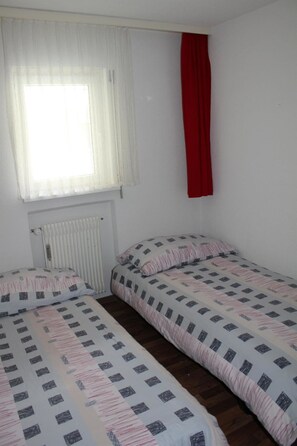 A room with two single beds, a window with sheer curtains, a radiator, and a red curtain on the wall. - Salute 206 # 4-bed apartment, Dusche, WC 4-Bettwohnung, Dusche, WC by Interhome (Leukerbad)