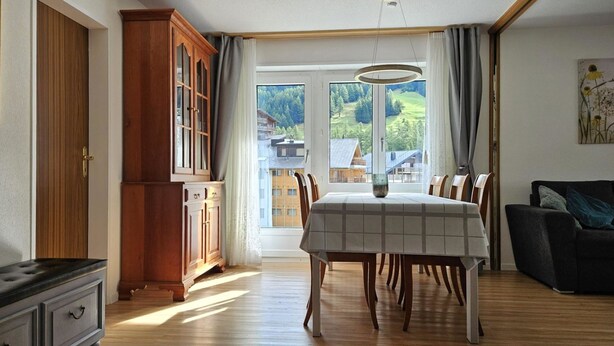 A dining area with a table and chairs, a wooden cabinet, and a view of a building through a window. - Europe 16 # 4-bed apartment, Bad, WC 4-bed apartment, Bad, WC by Interhome (Leukerbad)