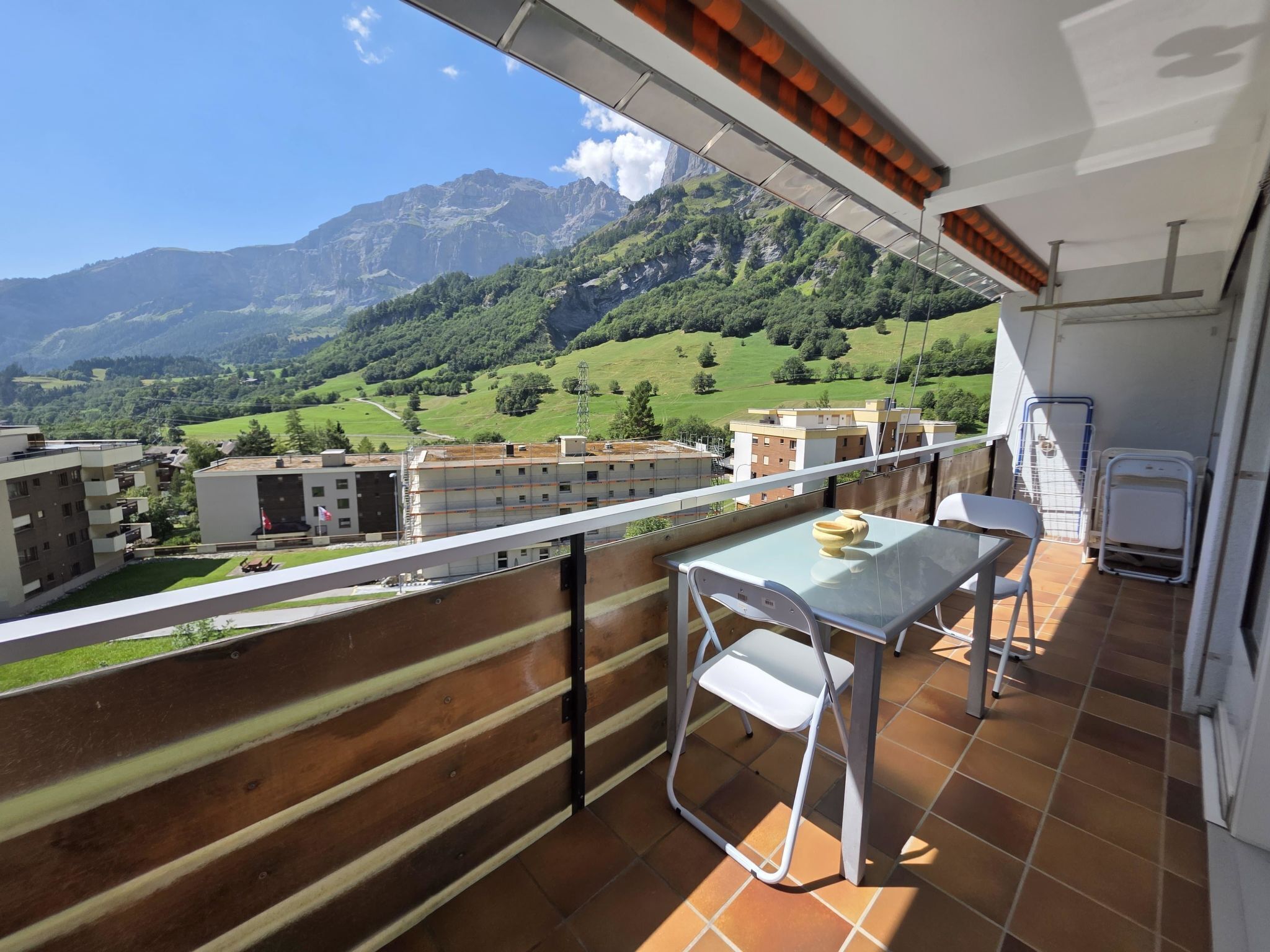 A balcony with a table and chairs, overlooking a mountainous landscape with buildings in the distance.
