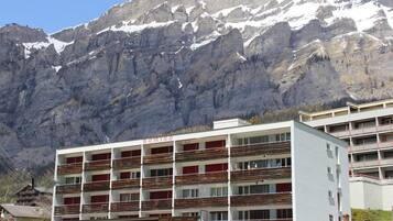 A multi-story hotel with balconies, a parking area, and a mountainous backdrop.