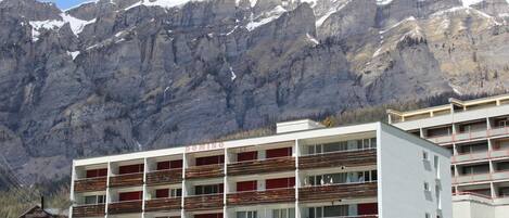 A multi-story hotel with balconies, a parking area, and a mountainous backdrop.
