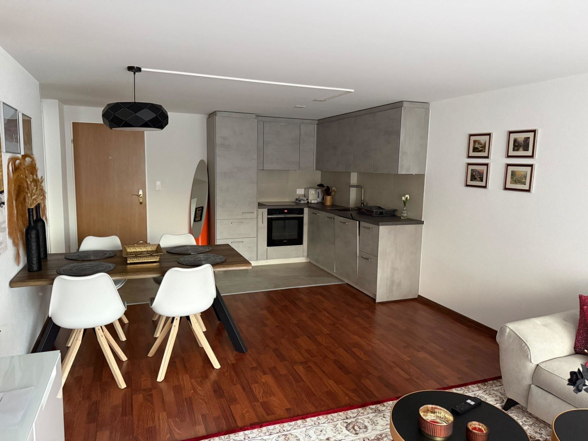 Eine moderne Küche mit grauen Schränken, ein Essbereich mit einem Holztisch und weißen Stühlen sowie ein Wohnbereich mit einer Couch und einem Couchtisch.