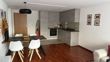 Eine moderne Küche mit grauen Schränken, ein Essbereich mit einem Holztisch und weißen Stühlen sowie ein Wohnbereich mit einer Couch und einem Couchtisch.