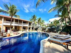 A swimming pool with lounge chairs, palm trees, and a building with balconies. - PortoFino (Porto Seguro)