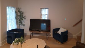 Un salon avec un téléviseur à écran plat, deux fauteuils bleus, une table basse ronde et une plante en pot.