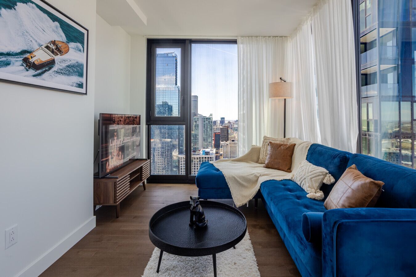 A modern living room with a blue sofa, a small round table, a television, and a large window with a city view.