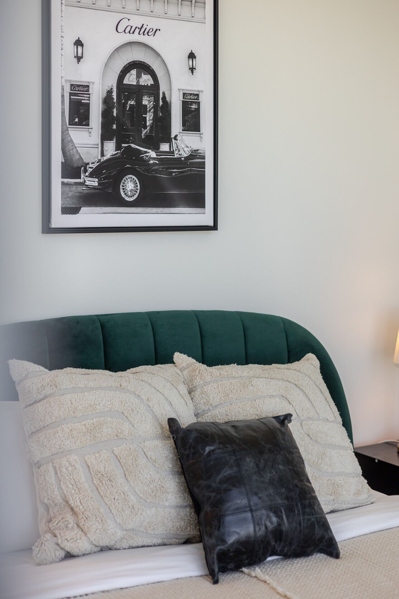A bed with a green upholstered headboard, two white textured pillows, and a black leather pillow. Above the bed, there is a framed black and white photograph of a car and storefront with the word 'Cartier' visible.