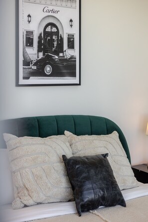 A bed with a green upholstered headboard, two white textured pillows, and a black leather pillow. Above the bed, there is a framed black and white photograph of a car and storefront with the word 'Cartier' visible. - The Azure Rise | 2B1B Condo in Downtown MTL (Montreal)