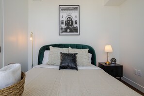 A neatly made bed with a green upholstered headboard, a framed picture above it, a bedside lamp, and a small clock on a nightstand. - The Azure Rise | 2B1B Condo in Downtown MTL (Montreal)
