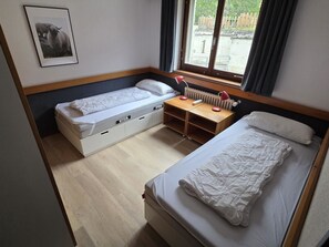 A room with two beds, a desk, a radiator, and a window with curtains. - Floralp 5 # 4-bed apartment, Bad, WC 4-Bettwohnung, Bad, WC by Interhome (Leukerbad)