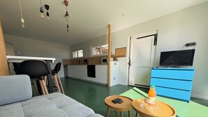 A modern living room with a grey sofa, two wooden side tables, a television, and a kitchen area with white cabinets and a blue cabinet.