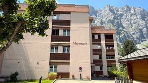 Un immeuble à plusieurs étages, portant l’inscription « Mayenzett » sur sa façade, entouré de verdure et avec des montagnes en arrière-plan.