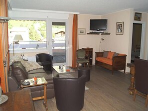 Un salon comprenant un canapé, un fauteuil et un pouf, un téléviseur fixé au mur et une fenêtre donnant sur un paysage verdoyant.