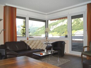 Ein Wohnzimmer mit einer Couch, Sesseln, einem Couchtisch und Blick auf eine gebirgige Landschaft durch große Fenster.
