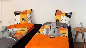 Una habitación con dos camas, cada una con una colcha y almohadas estampadas, una mesita de noche de madera y un televisor montado en la pared.