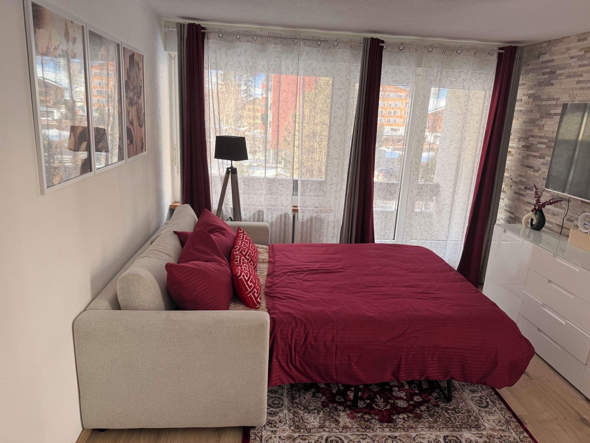 Un dormitorio con cama, sofá, lámpara y vistas a edificios a través de la ventana.