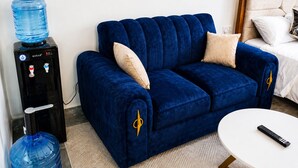 A blue sofa with a water dispenser, a remote control on a white table, and a bed with white bedding. - Modern, secure studio with Wi-Fi, Smart TV & kitchenette in Thindigua. (Kiambu)