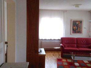 A living room with a red sofa, a small table, a radiator, and a window with curtains. - Royal 50 # 4-bed apartment, Dusche, WC 4-Bettwohnung, Dusche, WC by Interhome (Leukerbad)