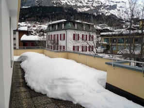 Een sneeuwbedekt balkon met uitzicht op een gebouw met rode luiken en een berg op de achtergrond.