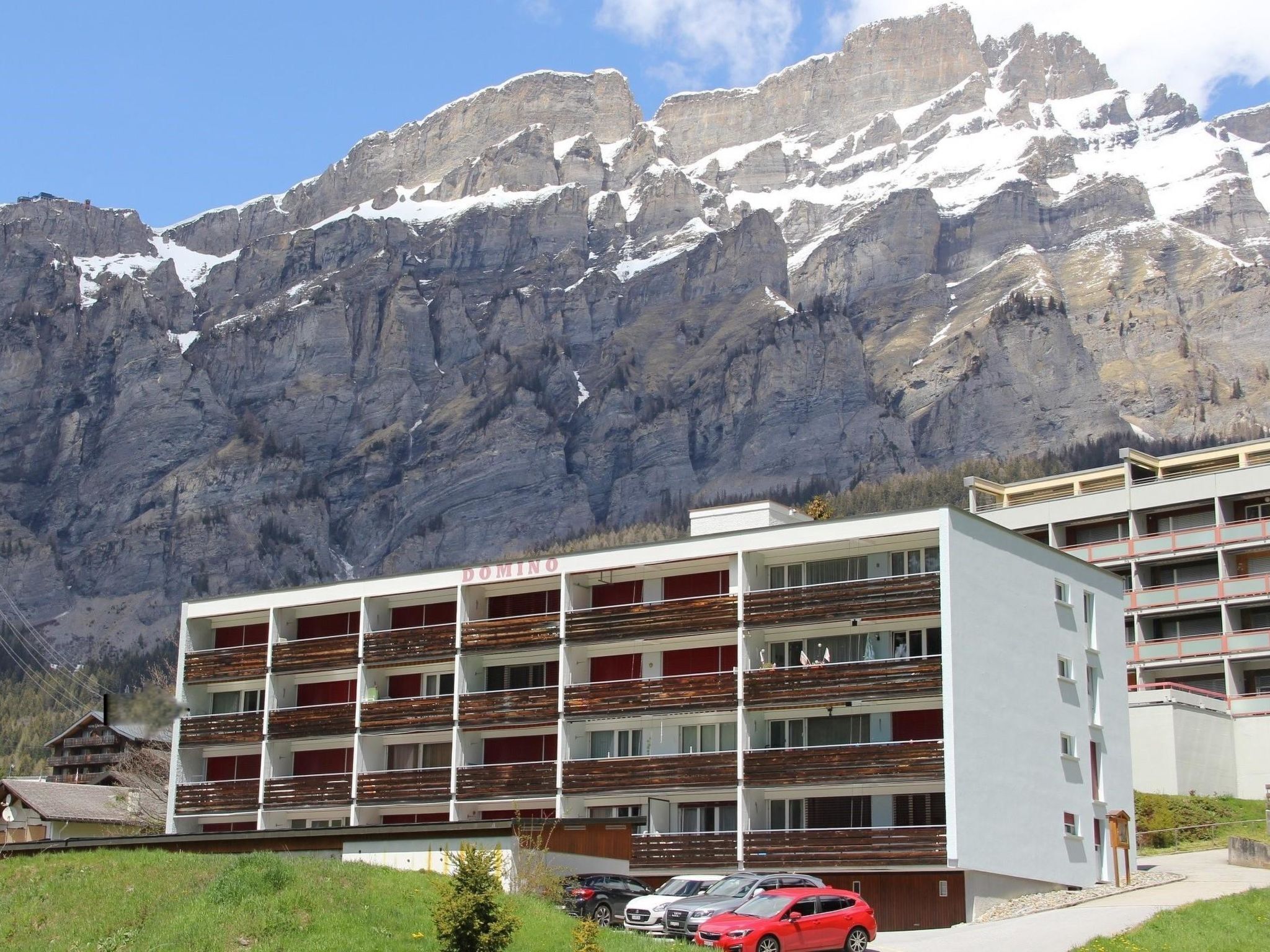 A multi-story building with balconies, a parking area, and a mountainous backdrop.