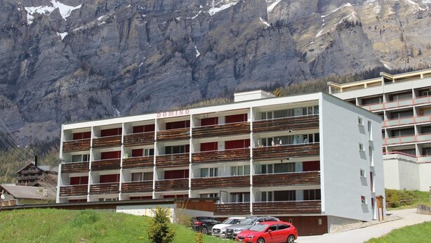Ein mehrstöckiges Gebäude mit Balkonen, einem Parkplatz und einer Bergkulisse.