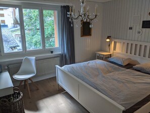 A bedroom with a bed, a chair, a bedside table, a lamp, a radiator, and a window with curtains.