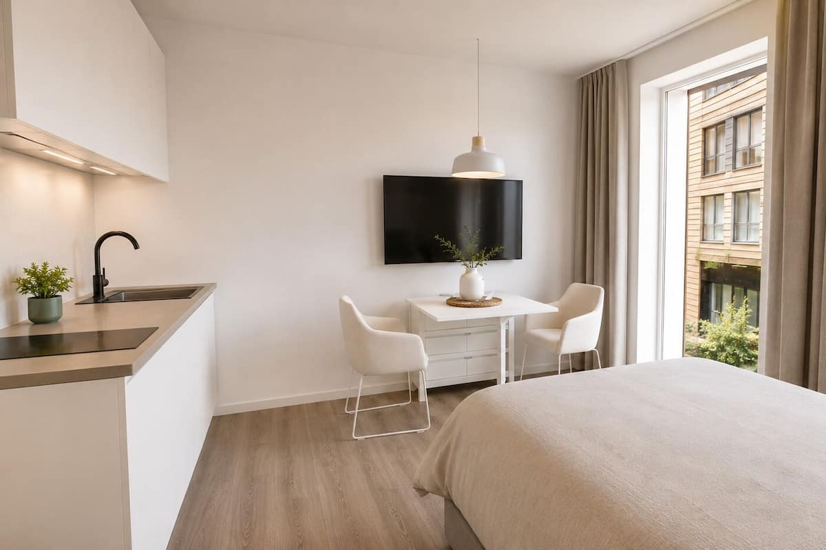A hotel room with a bed, a desk with two chairs, a TV, a potted plant, and a window with curtains.