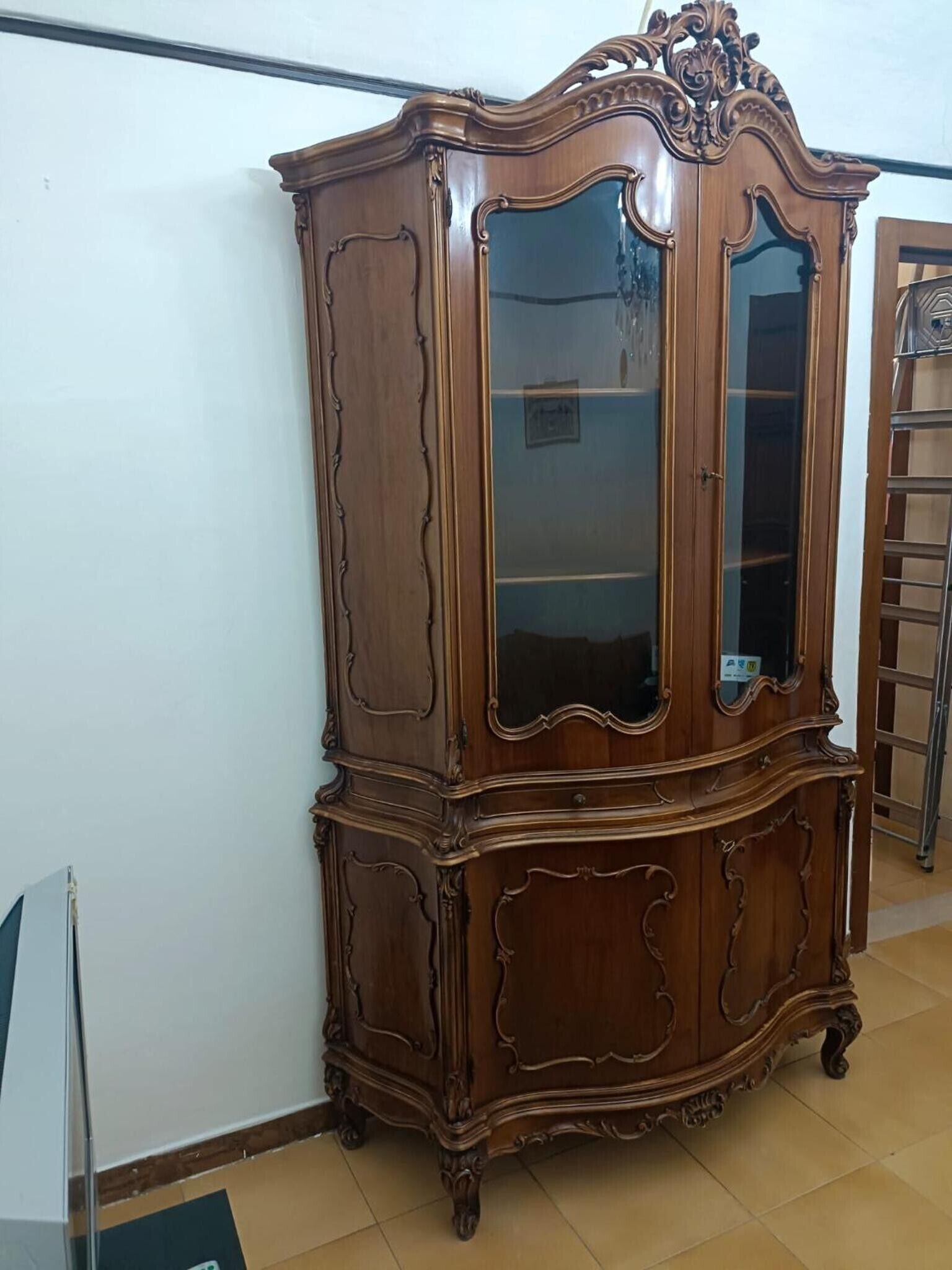 Une armoire en bois, dotée de portes en verre et de sculptures complexes, se trouve dans une pièce dont le sol est recouvert de carreaux et un des murs est peint en blanc.