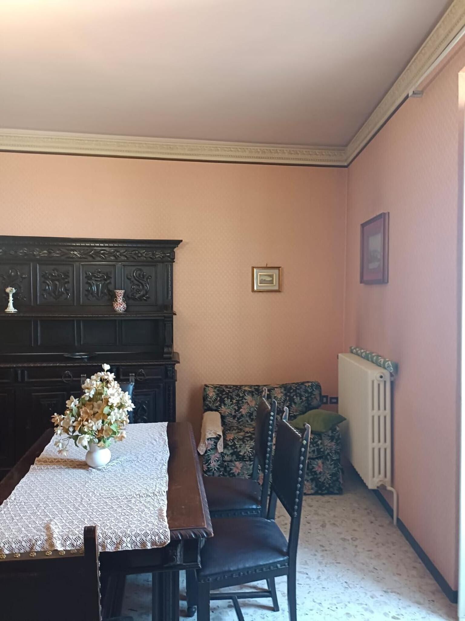 Un coin repas comprenant une table, des chaises, un buffet en bois foncé, un radiateur et un canapé recouvert d’un tissu à motifs floraux.