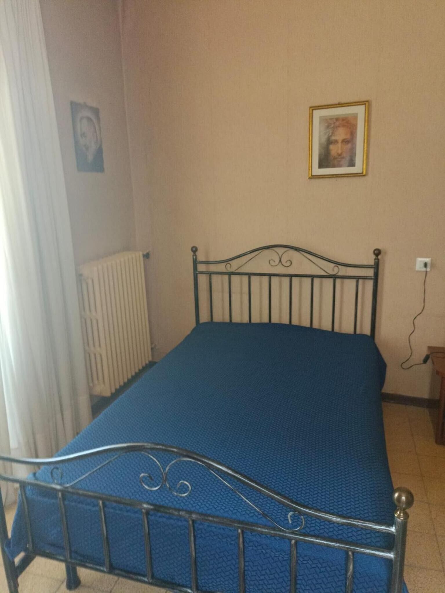 Ein Einzelbett mit Metallgestell und einer blauen Matratzenauflage in einem Zimmer mit Heizkörper und einem Wandbild.