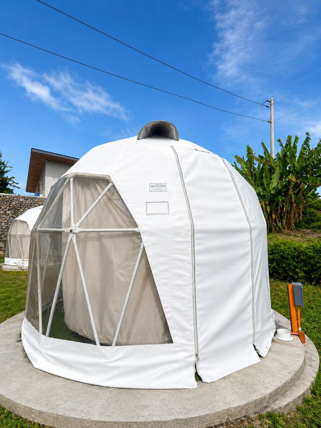A dome-shaped tent with a label 'MULTI-MAN' on the side, set on a concrete base outdoors.