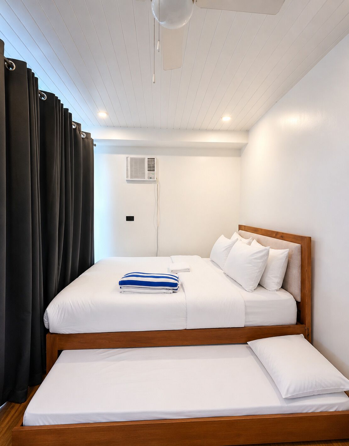 A bedroom with a double bed, a bedside table, a small bedside table, a wall-mounted air conditioning unit, and a window with curtains.
