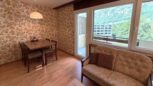 Ein Zimmer mit Esstisch, Stühlen, einer Couch und einem großen Fenster mit Blick auf ein Gebäude und Grünflächen.