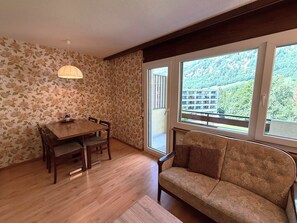 A room with a dining table, chairs, a sofa, and a large window with a view of a building and greenery. - Royal 33 # 2-bed apartment, Dusche, WC 2-Bettwohnung, Dusche, WC by Interhome (Leukerbad)