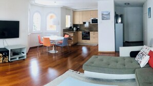 Un salon moderne avec un canapé gris, un coin repas avec des chaises orange et une cuisine dotée de meubles en bois.