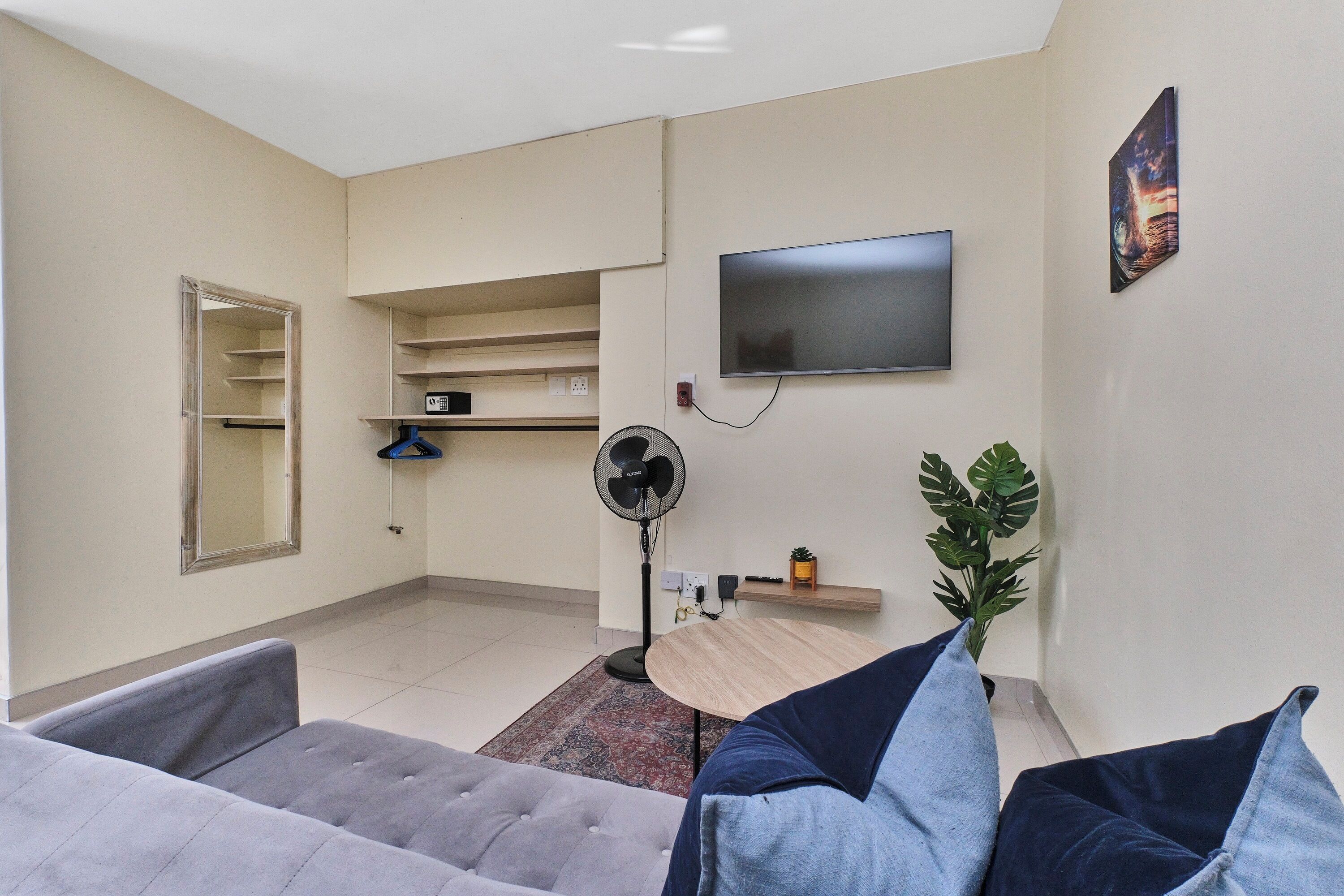 A living room with a grey sofa, a small table, a fan, a TV, and a plant.