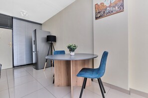 A modern dining area with a round table, blue chairs, a potted plant, and a wall-mounted lamp. - Urban Luxe Studio Illovo Mews by CTHA (Sandton)