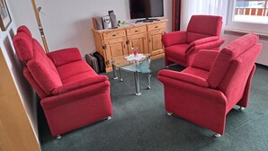 Una habitación con dos sillones rojos, una pequeña mesa de vidrio, un mueble de madera con un televisor y una ventana con vistas.