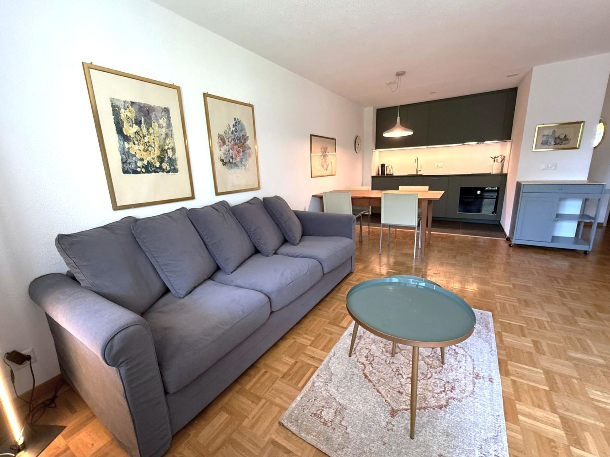 A modern living room with a grey sofa, a glass coffee table, and a kitchen in the background.