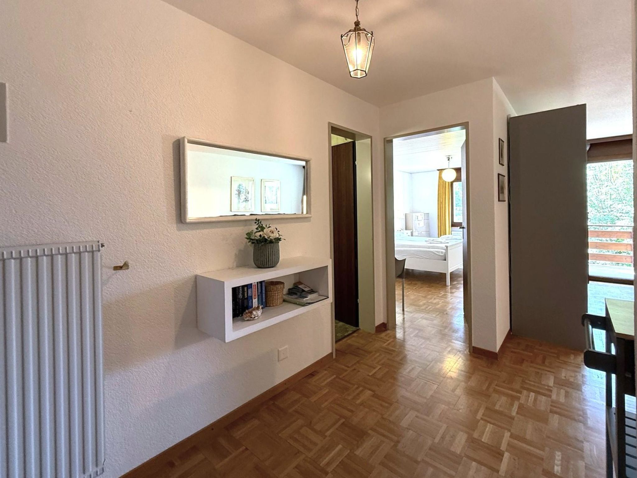 A hallway with a radiator, a shelf with books and a vase, and a door leading to a bedroom.