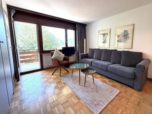 A living room with a grey sofa, two small side tables, a TV, and a large window with curtains. - Royal 41 # 4-bed apartment, Dusche, WC 4-Bettwohnung, Dusche, WC by Interhome (Leukerbad)