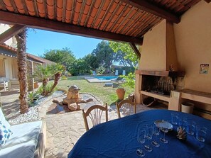 Uma área de estar coberta ao ar livre, com vista para a piscina, piso de pedra e churrasqueira.